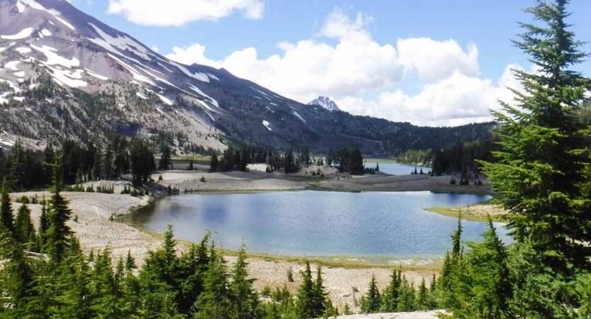 An alpine lake rests among evergreen trees and gray mountains dotted with snow